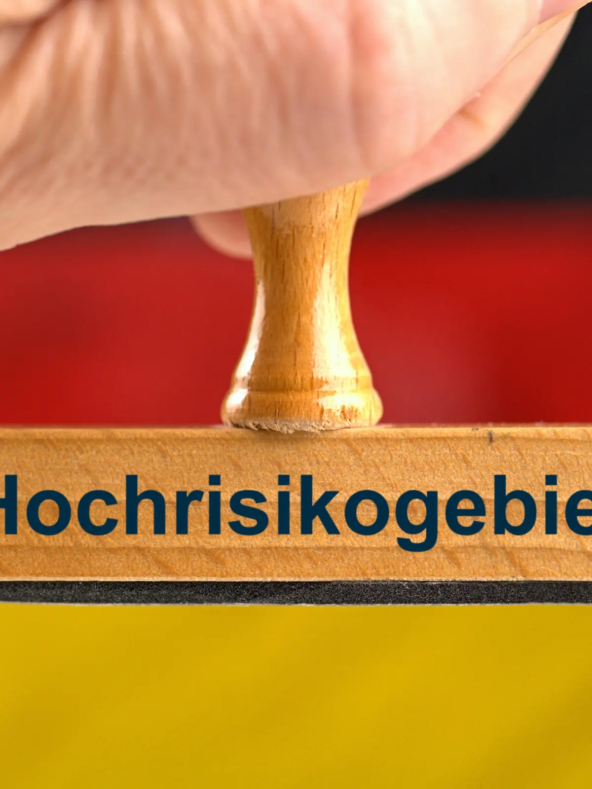 [M] Ein symbolischer Holzstempel mit der Aufschrift "Hochrisikogebiet", gehalten von einer Hand im Anschnitt vor einer unscharfen Deutschland-Flagge im Hintergrund. Composing als Symbolbild zur Frage, ob Deutschland im Ausland als Hochrisikogebiet eingestuft wird. || Modellfreigabe vorhanden