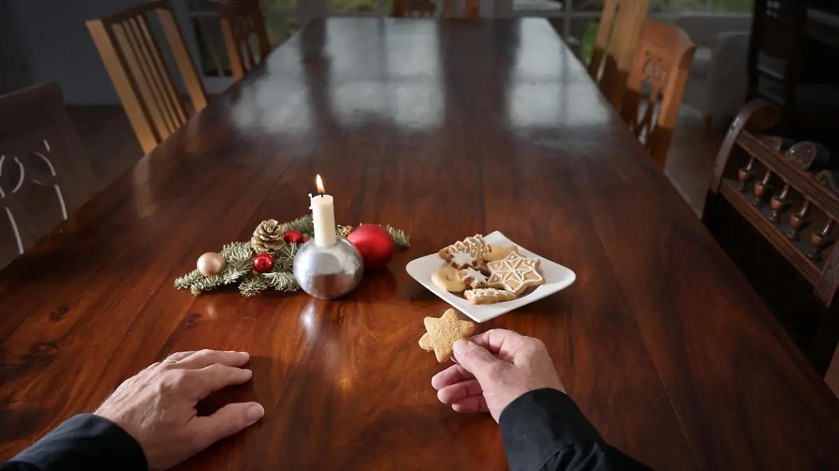 Hands of a lonely old man with a cookie sitting at a big family table with candle, Christmas decoration and empty chairs, holidays during the coronavirus pandemic, copy space, selected focus