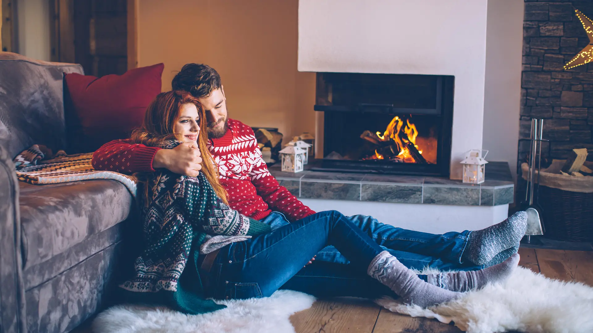Couple on vacation at mountain cabin. Sitting on the floor on a fur by fireplace in cozy  living room on Christmas. Wearing festive knitted sweaters and socks. Austrian Alps.