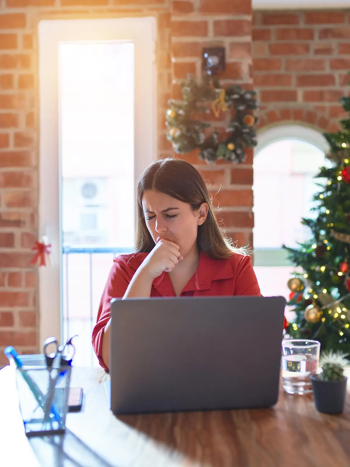 Beautiful woman sitting at the table working with laptop at home around christmas tree feeling unwell and coughing as symptom for cold or bronchitis. Healthcare concept.