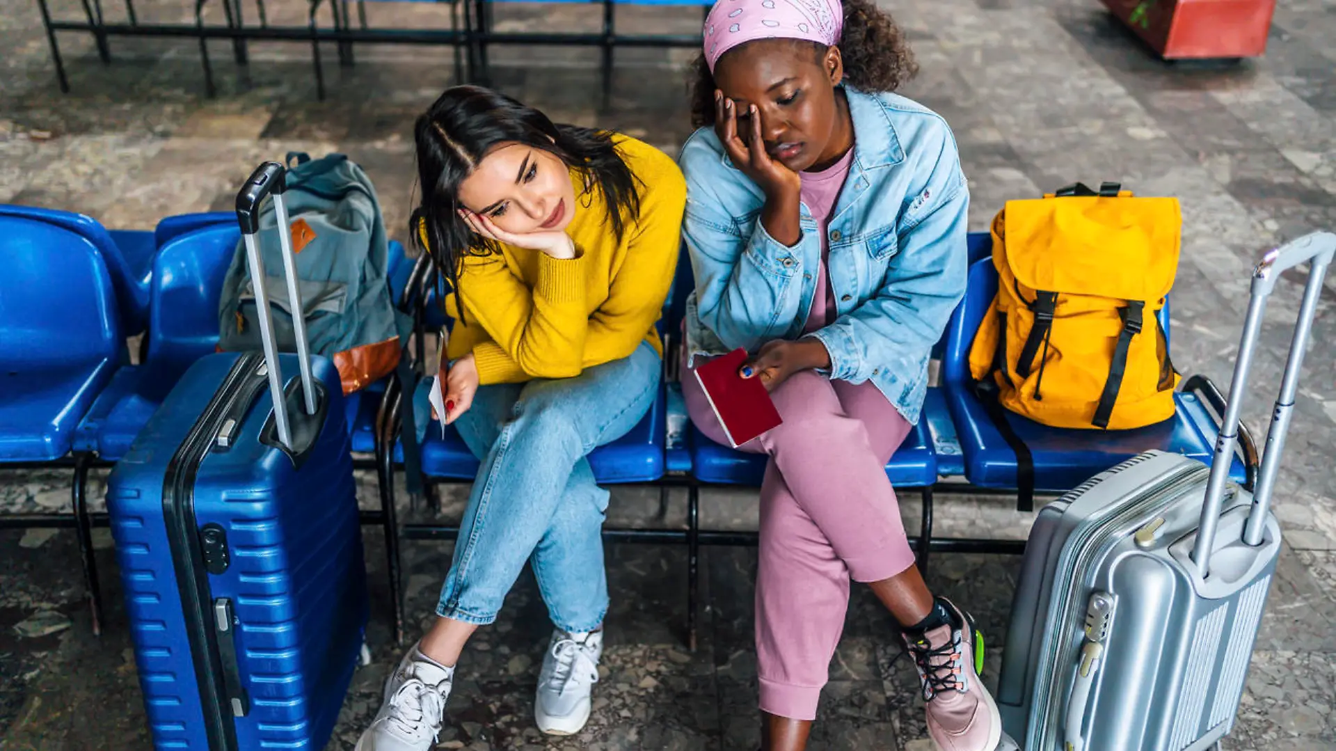Young multi racial women friends with suitcases sitting bored, waiting for a train at railway station