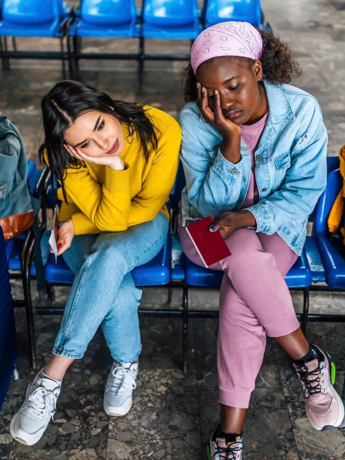 Young multi racial women friends with suitcases sitting bored, waiting for a train at railway station