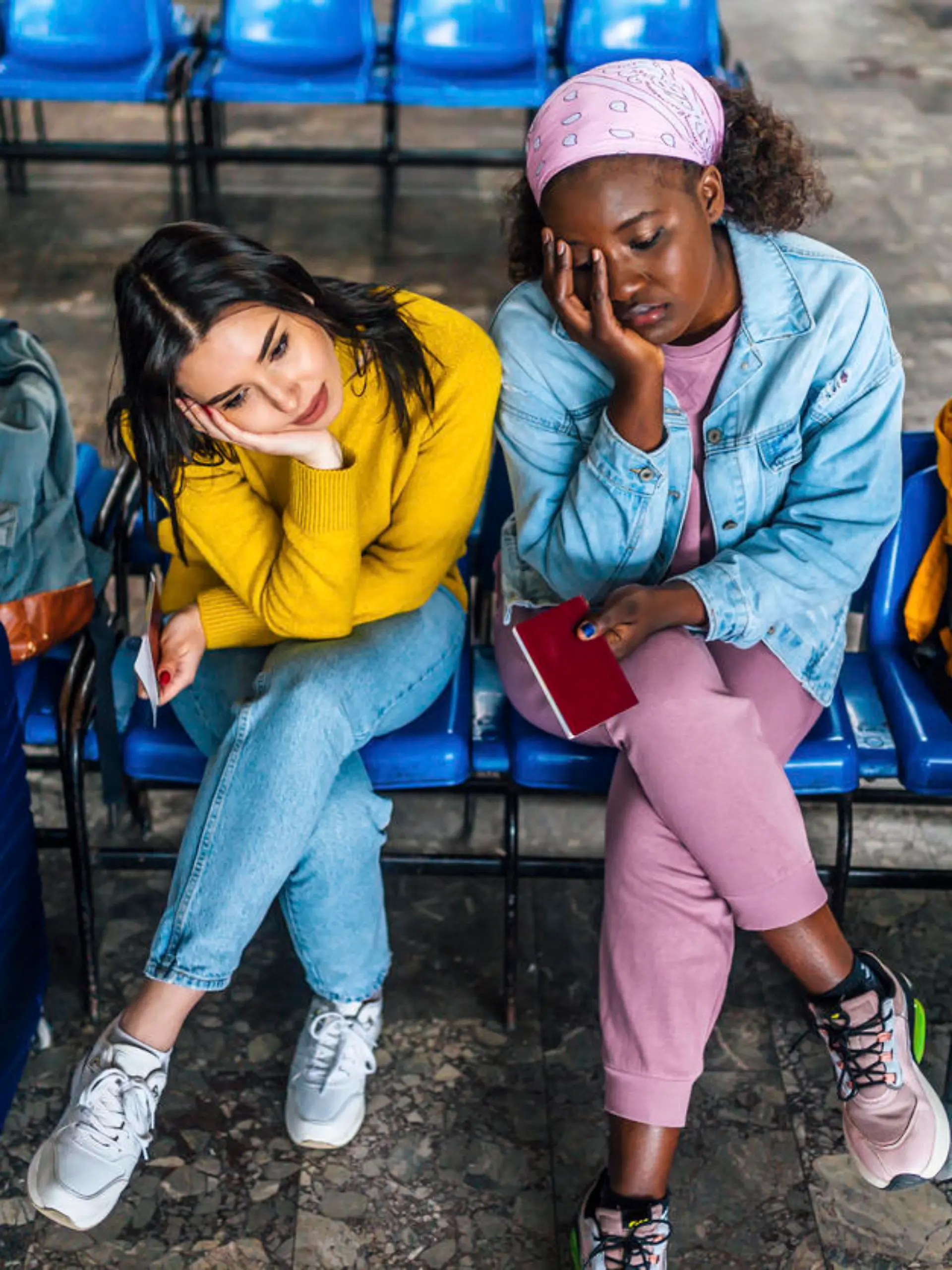 Young multi racial women friends with suitcases sitting bored, waiting for a train at railway station
