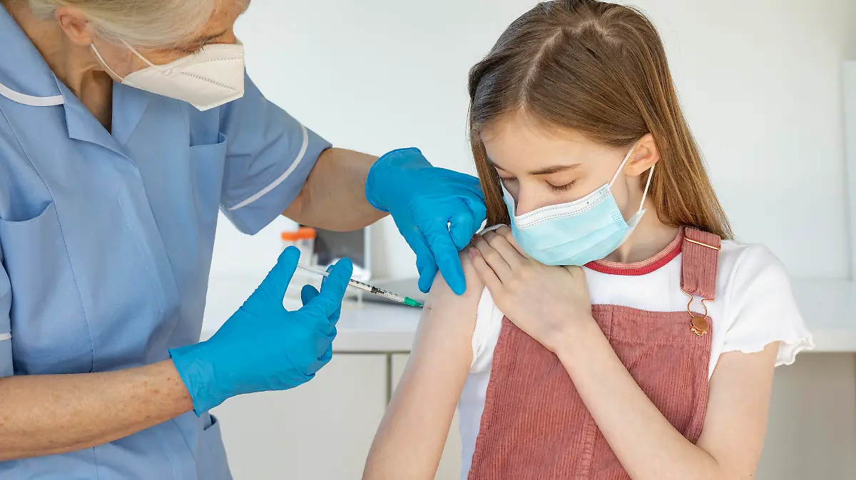 A medium shot of a Caucasian, young girl receiving her Covid-19 vaccination from a nurse. They are both wearing a protective face mask.