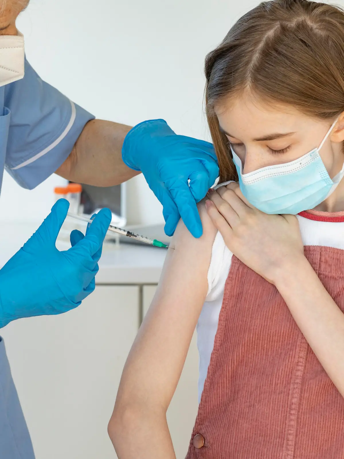 A medium shot of a Caucasian, young girl receiving her Covid-19 vaccination from a nurse. They are both wearing a protective face mask.