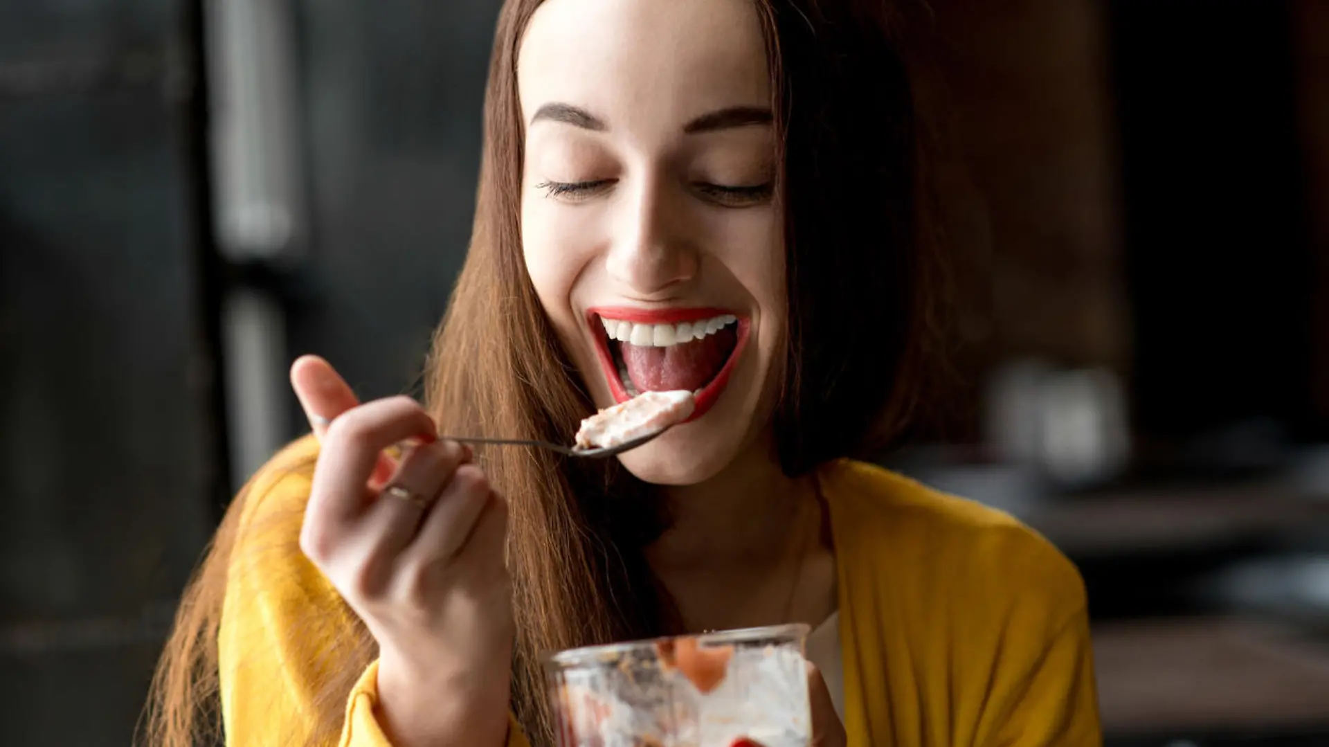 Young smiling woman dressed in yellow sweater eating ice cream in the dark cafe interior