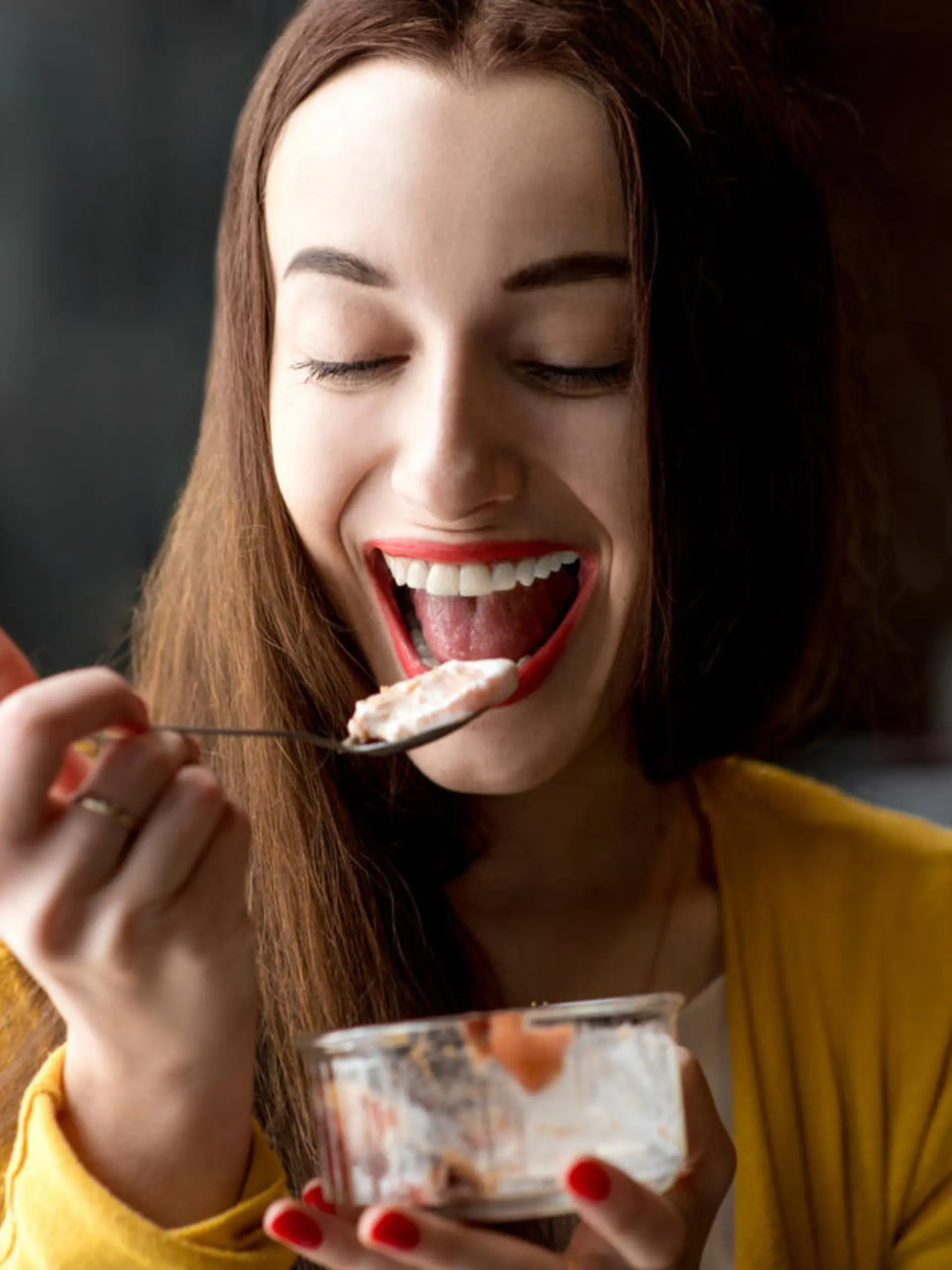 Young smiling woman dressed in yellow sweater eating ice cream in the dark cafe interior
