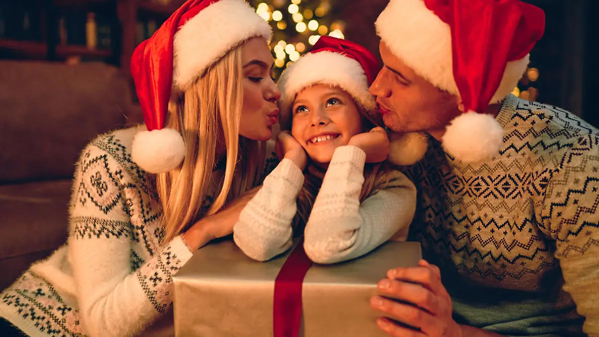 Merry Christmas and Happy New Year! Happy family is waiting for the New Year in Santa Claus hats exchange gifts with each other. Mom and dad kiss a cute daughter