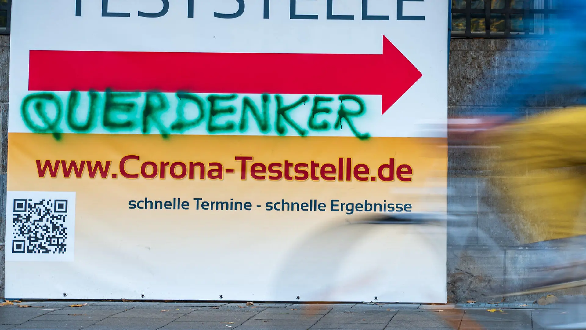 ARCHIV - 02.11.2021, Bayern, München: Ein Mann radelt an einem Plakat vorbei, auf das jemand das Wort «Querdenker» gesprüht hat. «Querdenker» und andere Begriffe im Zusammenhang mit der Corona-Pandemie dominieren auch die Suche nach dem Unwort des Jahres. (Illustration zu dpa: «Corona-Pandemie dominiert «Unwort des Jahres»-Suche») Foto: Peter Kneffel/dpa +++ dpa-Bildfunk +++