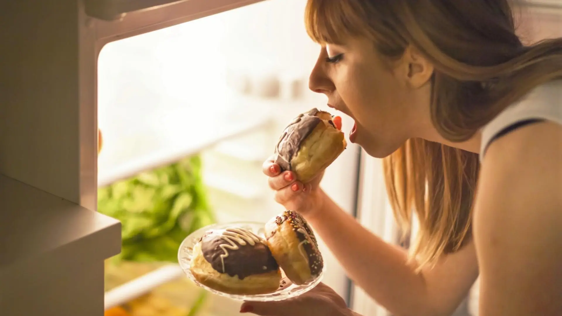 Close up image of a young woman with eating disorder, having a midnight snack - eating donuts, in front of the refrigerator.