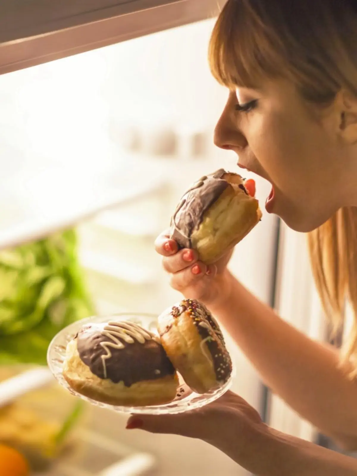 Close up image of a young woman with eating disorder, having a midnight snack - eating donuts, in front of the refrigerator.