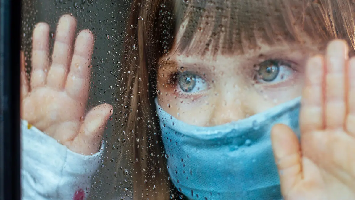 Little girl in face mask looking in the window glass with rain drops. Selective focus on the eyes. Social isolation stay at home during Pandemic COVID-19 concept.