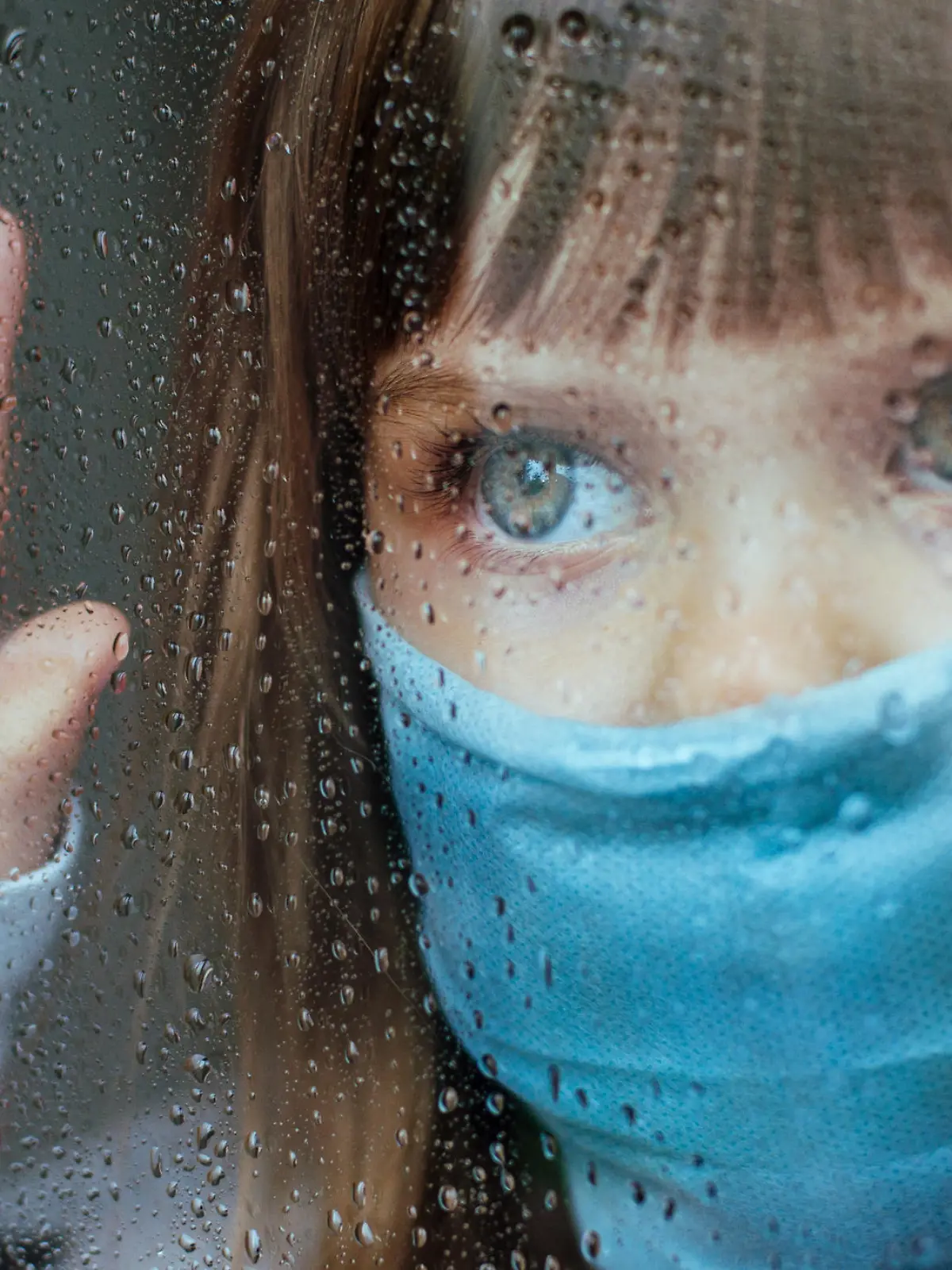 Little girl in face mask looking in the window glass with rain drops. Selective focus on the eyes. Social isolation stay at home during Pandemic COVID-19 concept.