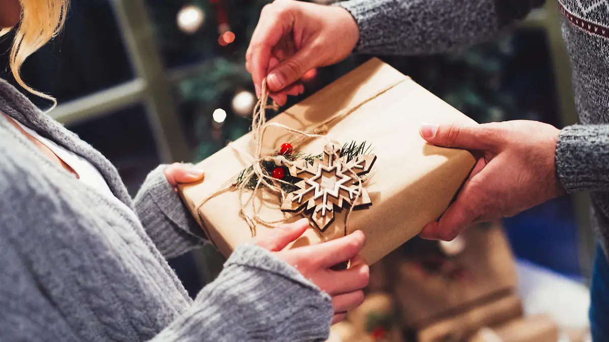A close-up shot of a man receiving a Christmas gift from a woman. The present is wrapped in kraft paper, decorated with a wooden snowflake and a peace of a pine tree branch and tied with manila rope. Using natural and recyclable materials is one of the ways to make your Christmas environmentally friendly.
