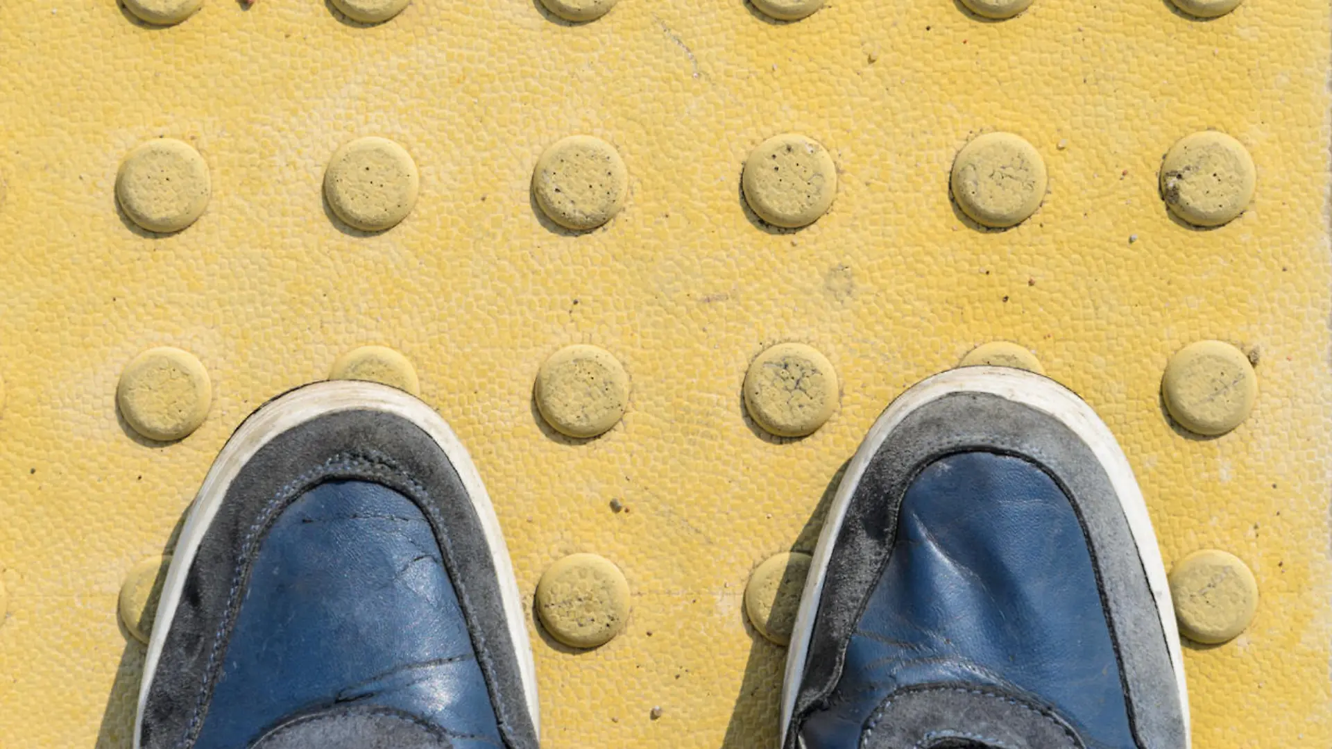 View from the First Person to his Feet, Standing in Front of the Sidewalk with Tactile Tiles to Navigate Blind People. Blue Sneakers on Yellow Tactile Paving Slabs.