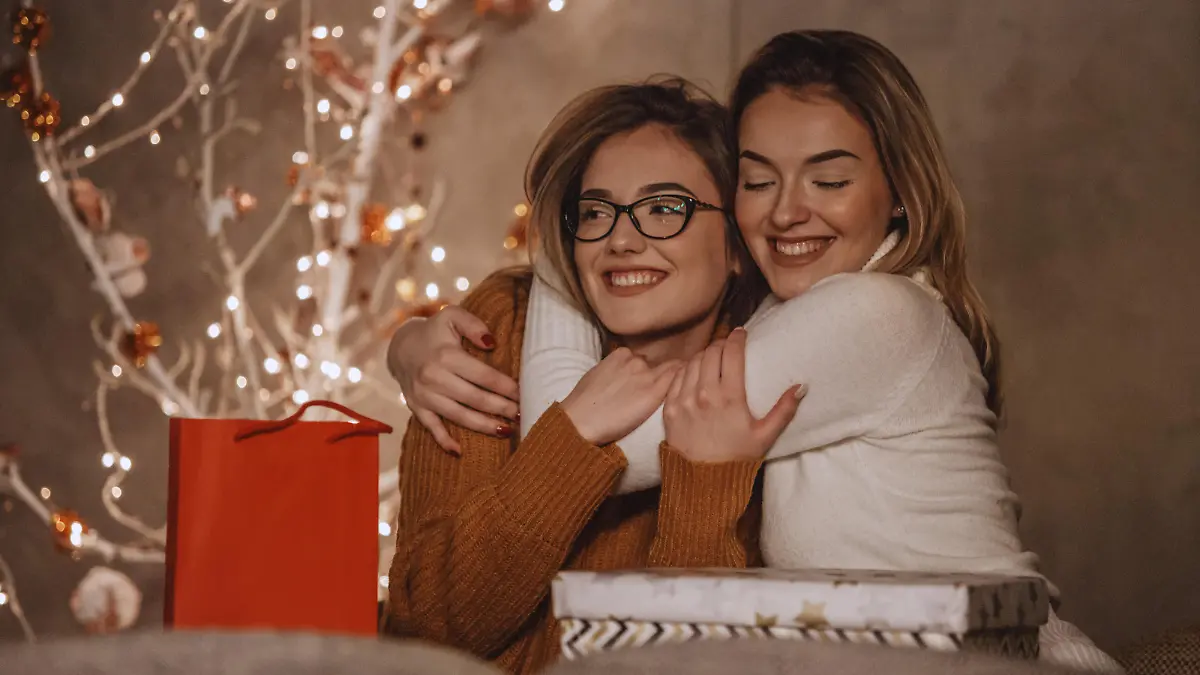 Two beautiful young women are hugging after exchanging gifts near the modern Christmas tree.