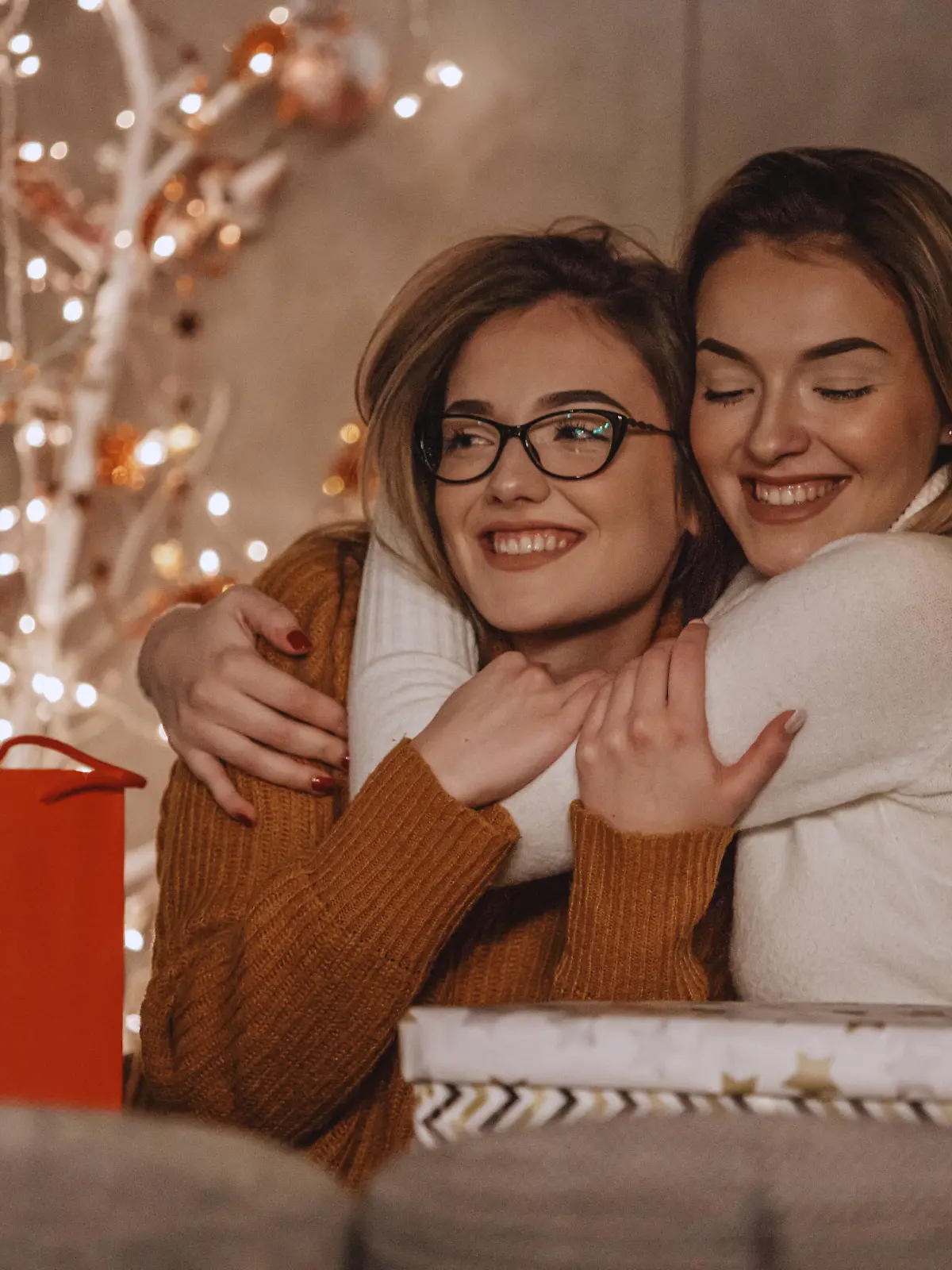 Two beautiful young women are hugging after exchanging gifts near the modern Christmas tree.