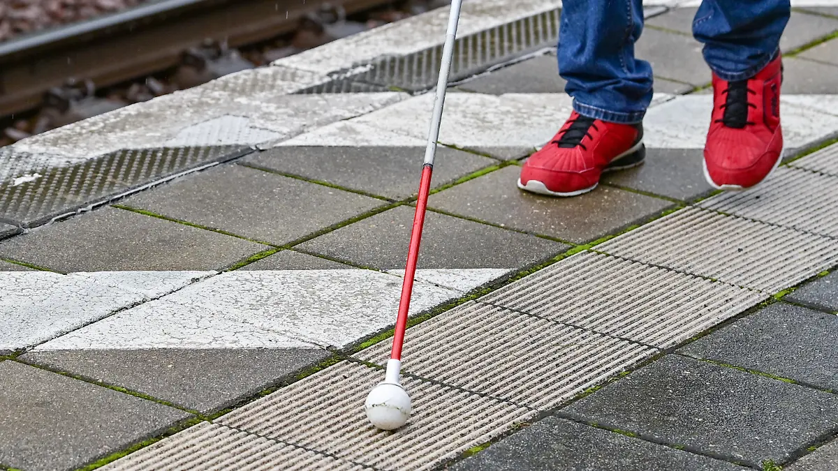 Ein Blindenstock mit einer Kugel ist auf einen Bahnsteig mit speziellen Bodenleitsystemen, auch Blindenleitsysteme genannt, zu sehen.