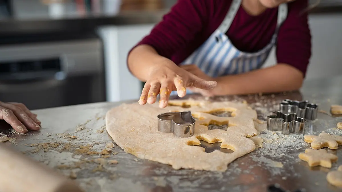 Closeup of child hands cutting cookies from dough. Little girl in apron pressing cookie cutter for shaping dough. Girl baking heart shaped christmas biscuits in the kitchen.