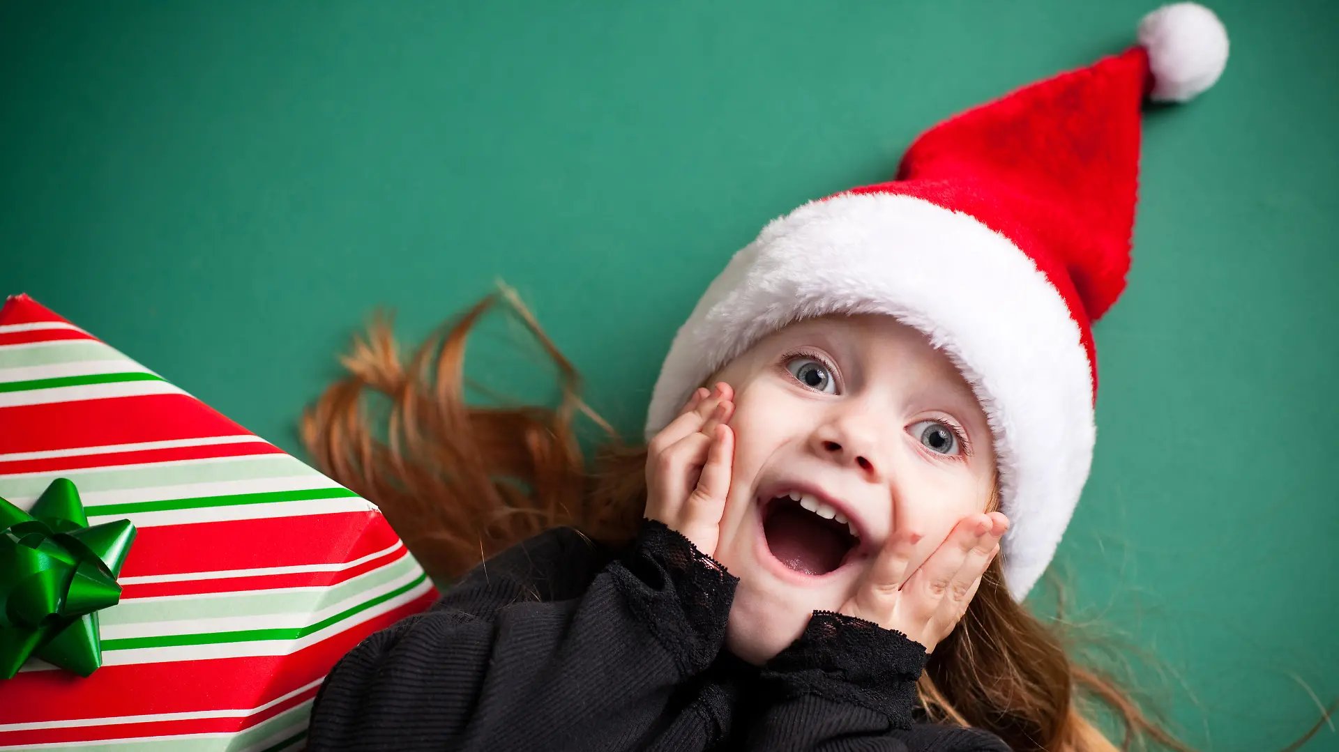 Color image of an excited 4-year-old girl wearing a Santa hat and lying on a green background.