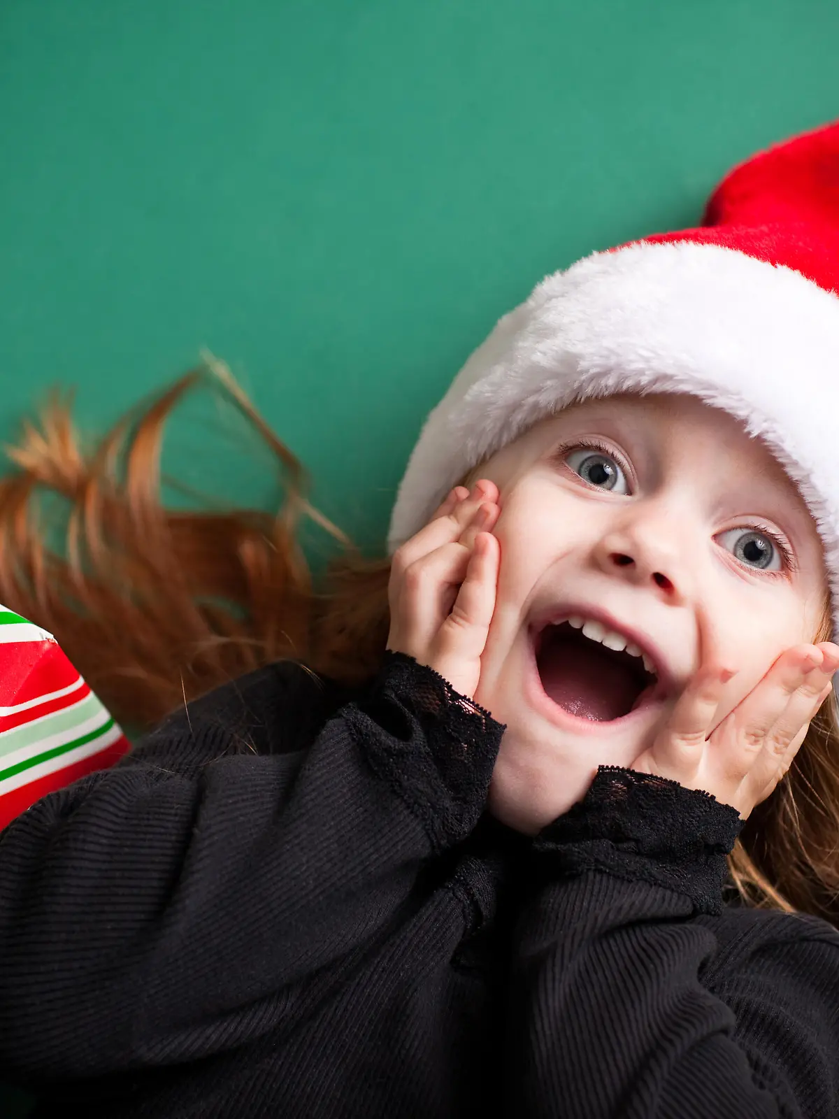Color image of an excited 4-year-old girl wearing a Santa hat and lying on a green background.