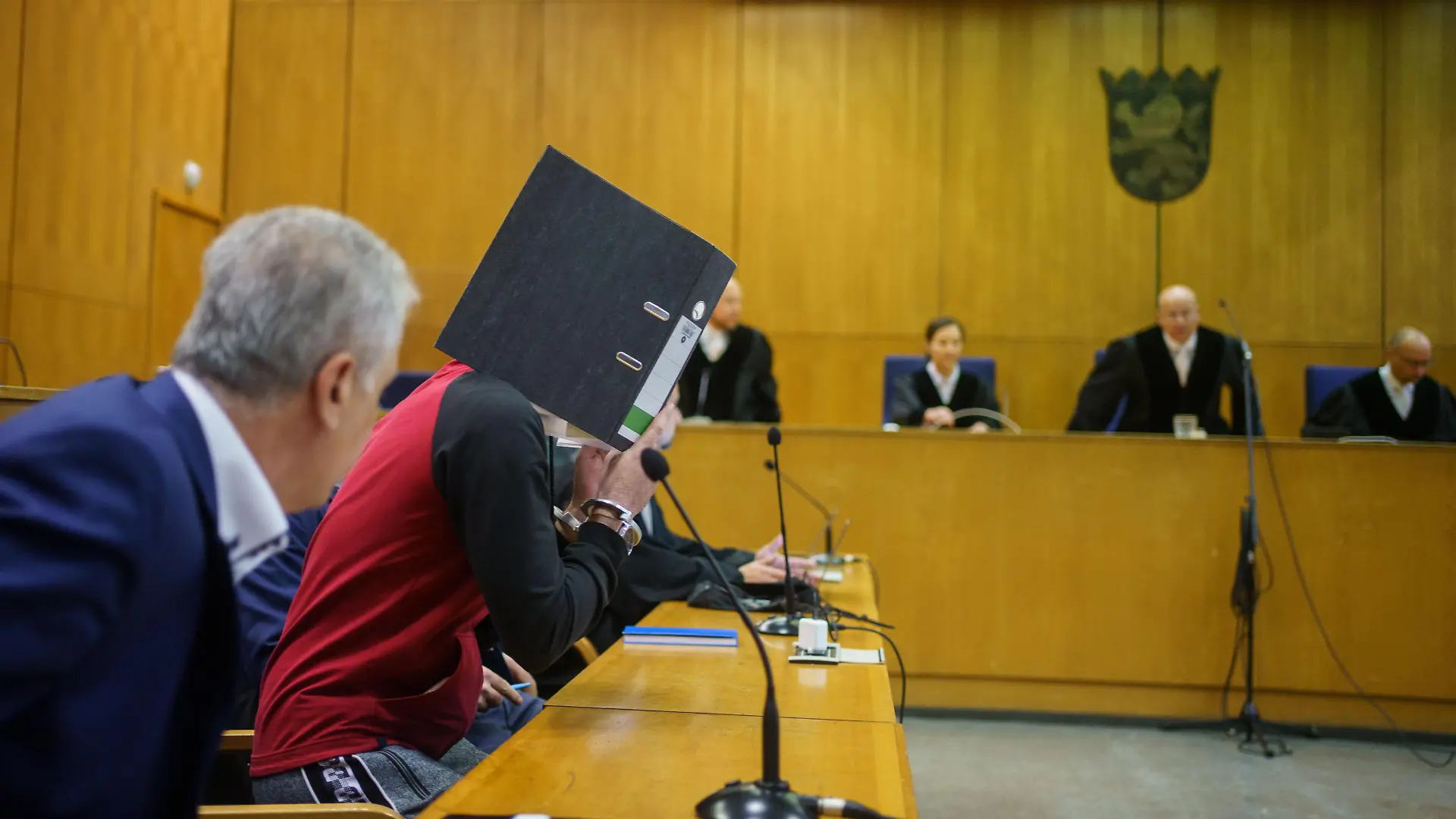 Mit einem Aktenordner vor dem Gesicht nimmt der Iraker Taha Al-J. (2.v.l) vor der Urteilsverkündung im Frankfurter Oberlandesgericht auf der Anklagebank Platz. Die Bundesanwaltschaft wirft ihm Völkermord, Verbrechen gegen die Menschlichkeit, Kriegsverbrechen, Menschenhandel und Mord vor. Als mutmaßliches Mitglied der Terrormiliz IS soll er eine Jesidin und ihre Tochter als Sklavinnen gehalten und misshandelt haben. Dabei soll er die Fünfjährige im irakischen Falludscha gefesselt haben, wo das Kind laut Anklage qualvoll verdurstete.