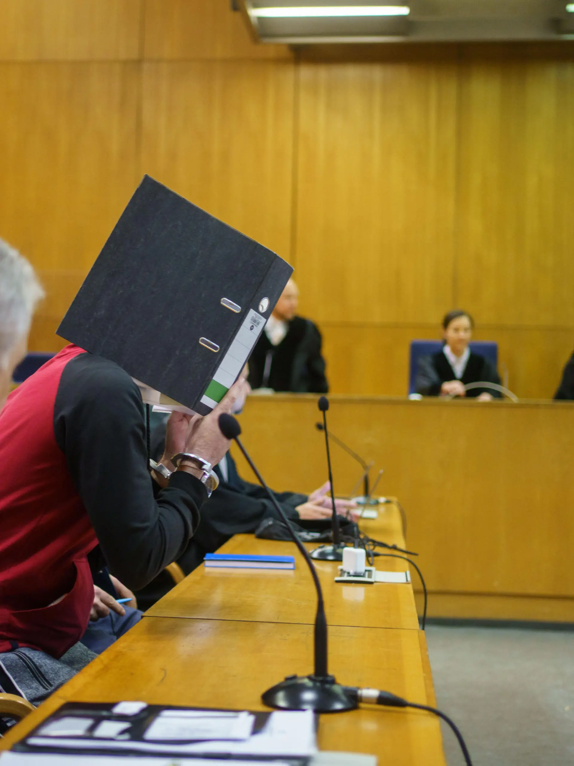 Mit einem Aktenordner vor dem Gesicht nimmt der Iraker Taha Al-J. (2.v.l) vor der Urteilsverkündung im Frankfurter Oberlandesgericht auf der Anklagebank Platz. Die Bundesanwaltschaft wirft ihm Völkermord, Verbrechen gegen die Menschlichkeit, Kriegsverbrechen, Menschenhandel und Mord vor. Als mutmaßliches Mitglied der Terrormiliz IS soll er eine Jesidin und ihre Tochter als Sklavinnen gehalten und misshandelt haben. Dabei soll er die Fünfjährige im irakischen Falludscha gefesselt haben, wo das Kind laut Anklage qualvoll verdurstete.