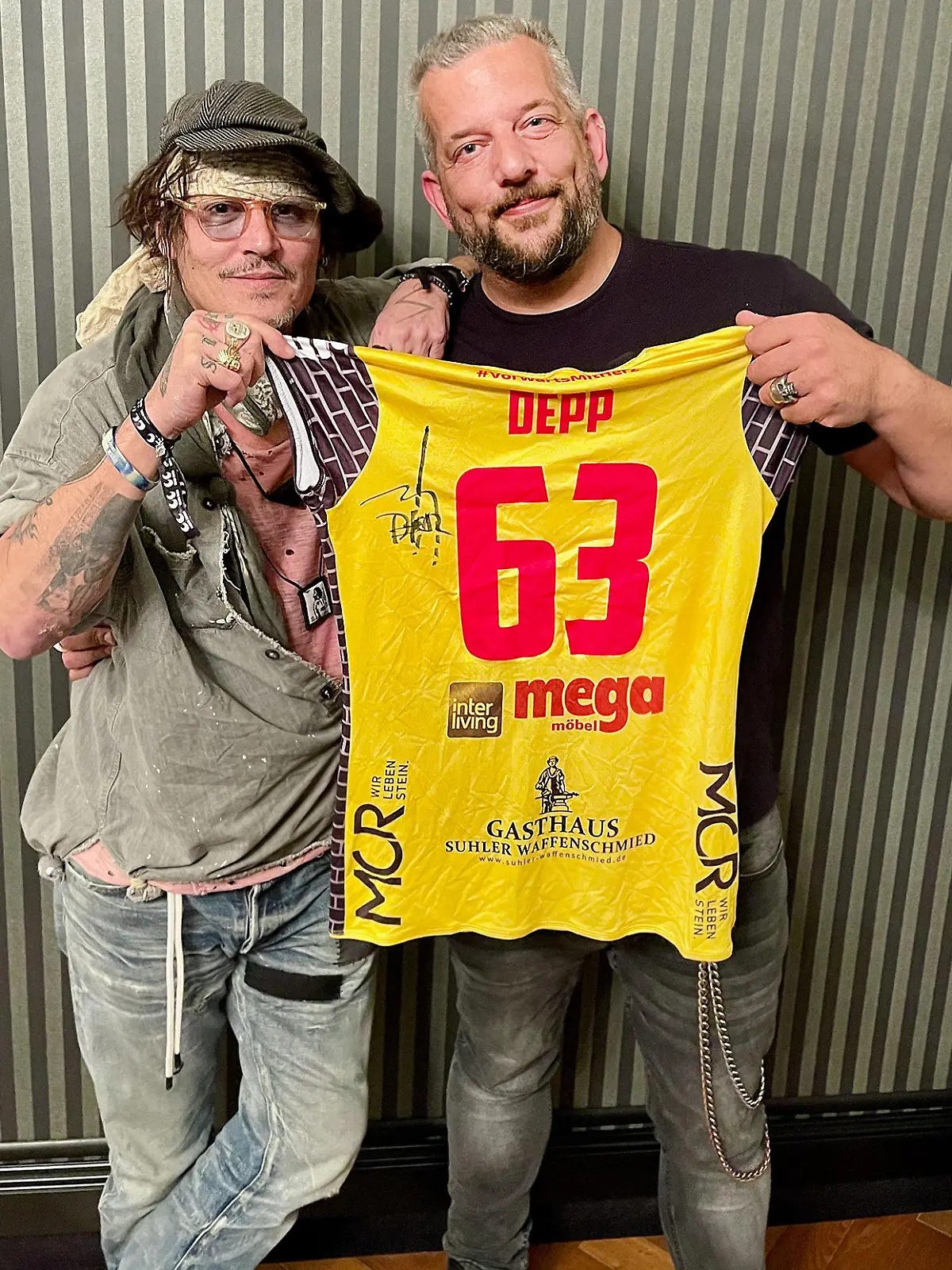 HANDOUT - 30.08.2021, Tschechien, Karlsbad: Johnny Depp (l) und der in Suhl lebende Musiker Alexander Becker halten zusammen ein Trikot des Volleyball-Bundesligistes VfB Suhl. Depp hatte das Trikot der VfB-Damen im August beim Filmfestival im tschechischen Karlsbad signiert, den Kontakt hatte Becker hergestellt. Im Oktober soll es versteigert werden, um den Verein zu unterstützen, teilte der VfB Suhl am Mittwoch mit. Foto: Johnny Depp : Alex Becker/VfB Suhl/dpa - ACHTUNG: Nur zur redaktionellen Verwendung im Zusammenhang mit der aktuellen Berichterstattung und nur mit vollständiger Nennung des vorstehenden Credits +++ dpa-Bildfunk +++