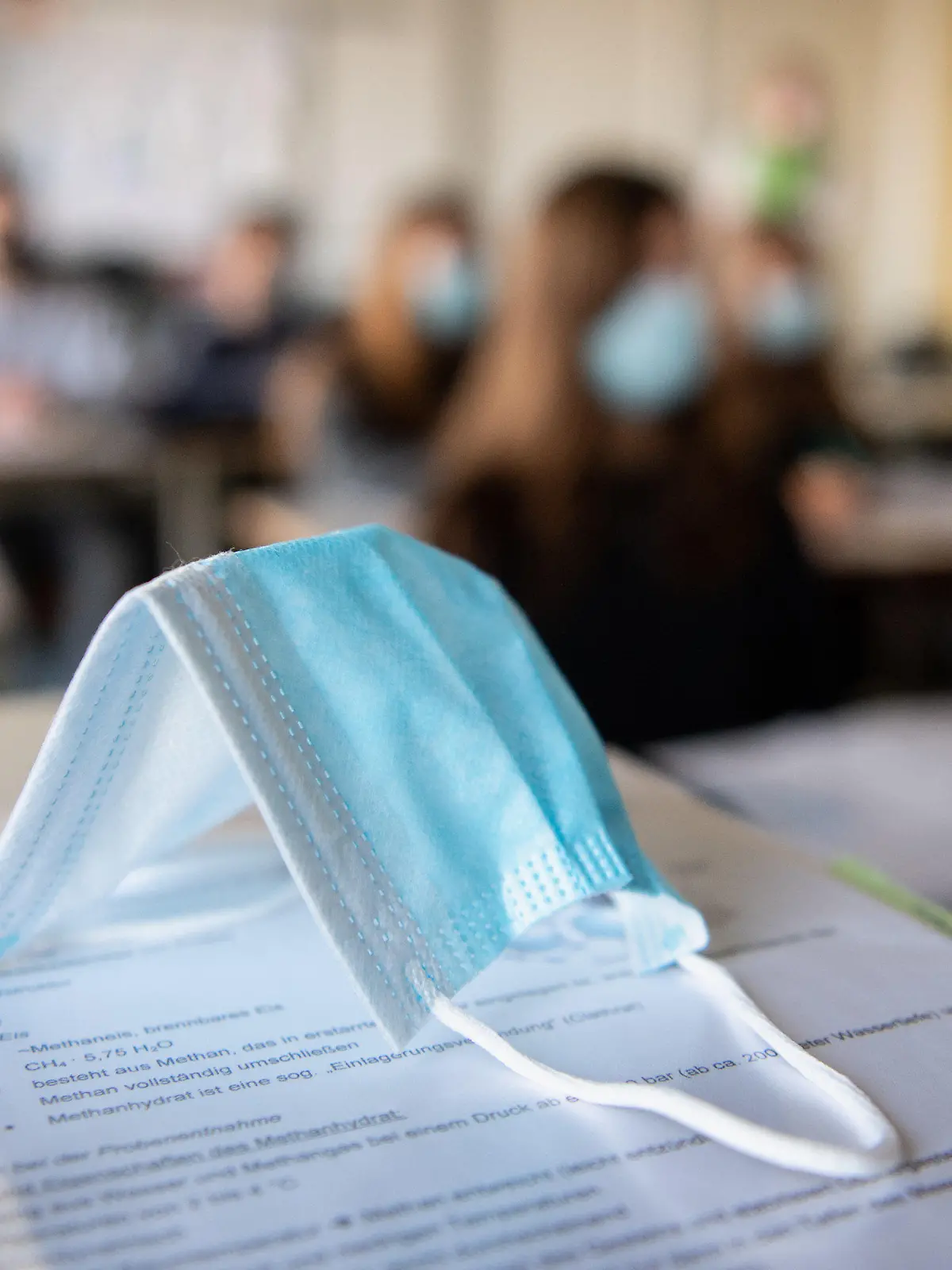 ARCHIV - 17.11.2020, Bayern, München: Eine Maske liegt im Unterricht auf Unterlagen, während im Hintergrund Schülerinnen und Schüler in einem Gymnasium mit Mund- und Nasenschutz zu sehen sind (zu dpa «Zurück ins Klassenzimmer - Weitere Schüler gehen wieder in die Schule») Foto: Matthias Balk/dpa +++ dpa-Bildfunk +++