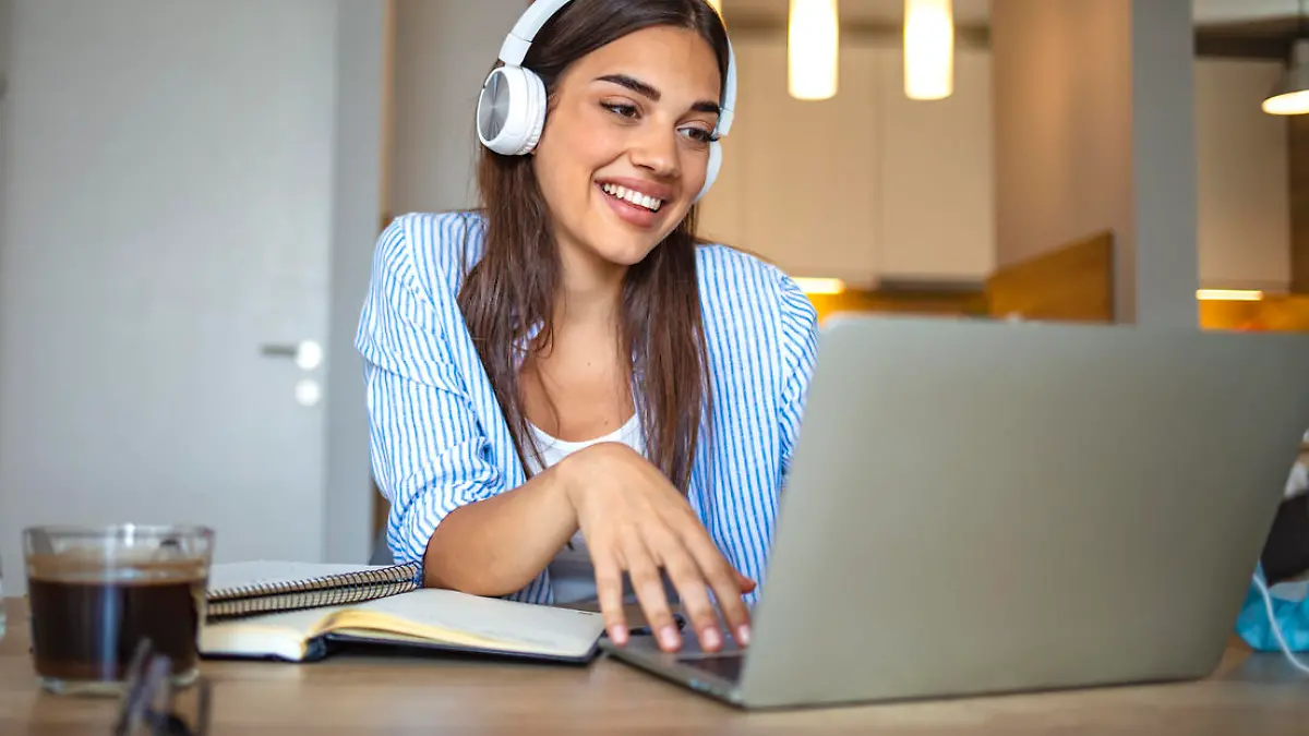 Smiling girl student wear wireless headphone study online with skype teacher, happy young woman learn language listen lecture watch webinar write notes look at laptop sit in cafe, distant education