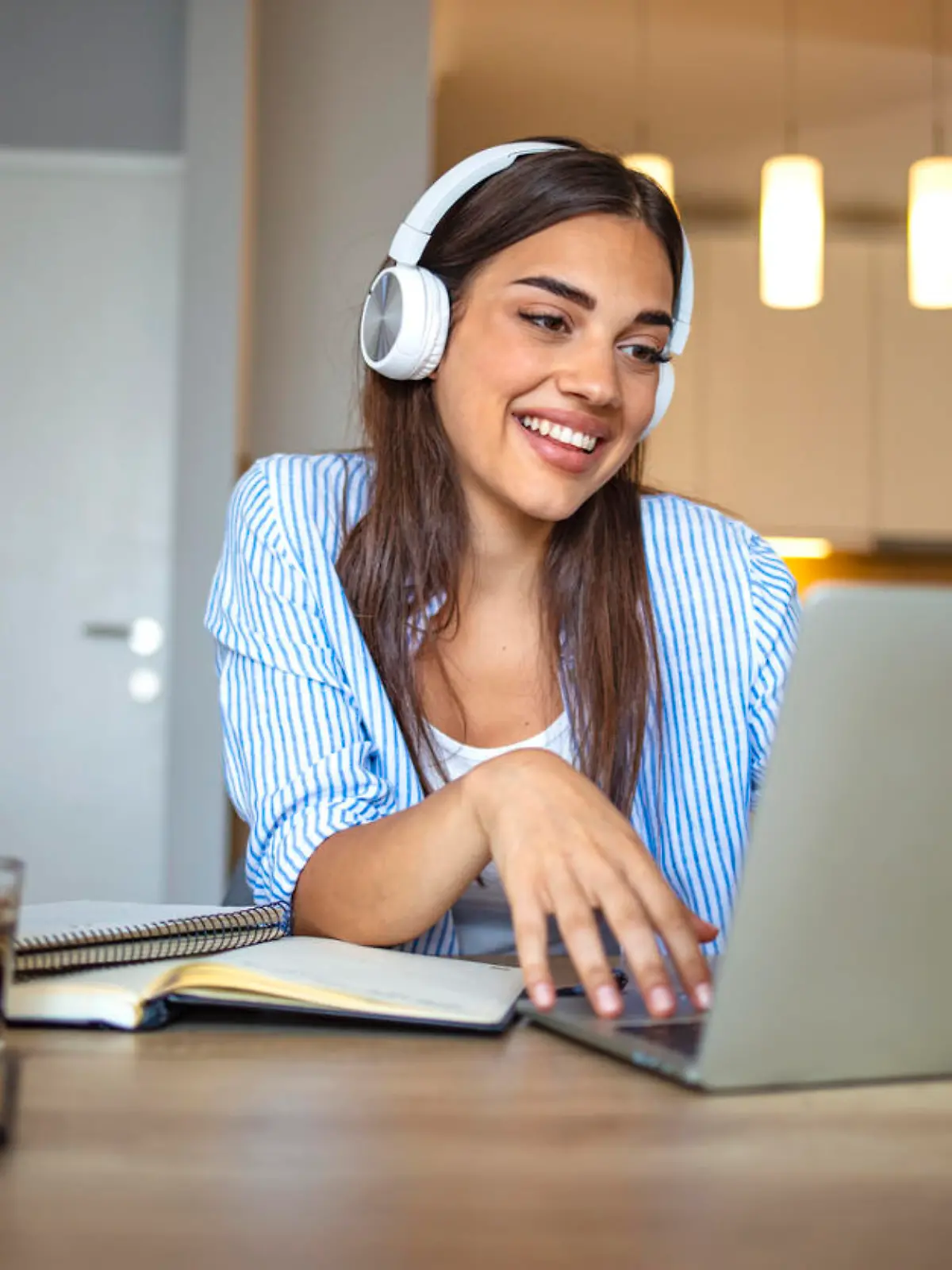 Smiling girl student wear wireless headphone study online with skype teacher, happy young woman learn language listen lecture watch webinar write notes look at laptop sit in cafe, distant education