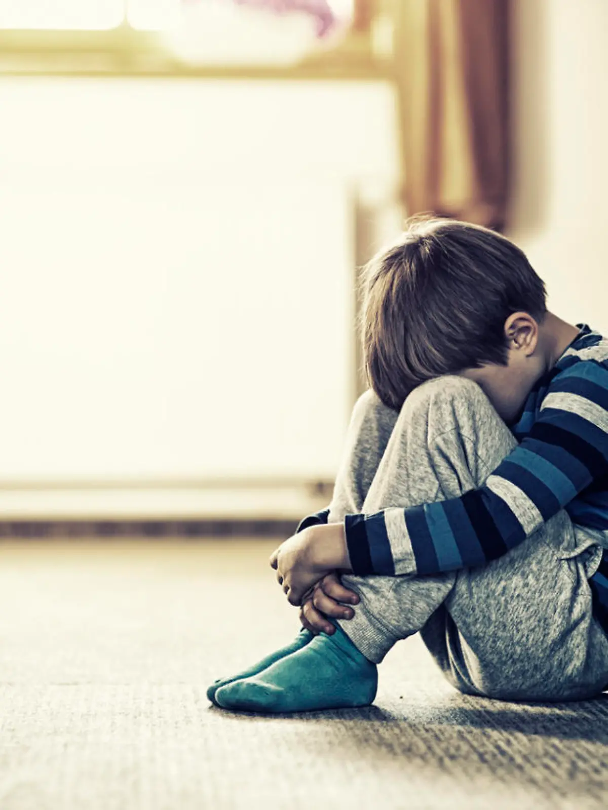 Depressed sad child sitting on the floor, in the door. The little boy is hiding his head between legs.