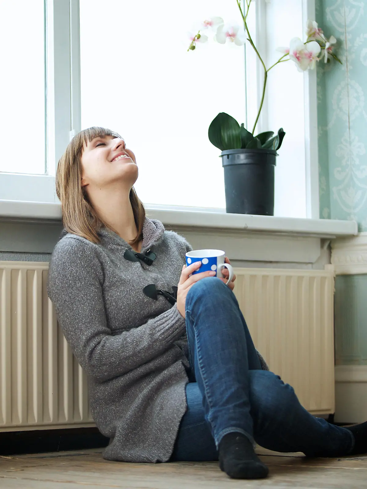 Portrait of a happy young woman sitting on the floor and leaning against radiator. Smiling female looking up and relaxing at home.