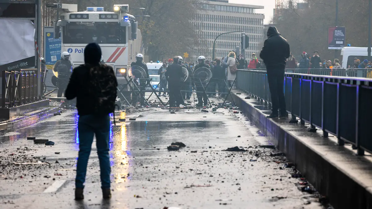 21.11.2021, Belgien, Brüssel: Steine und zersplitterte Flaschen liegen auf einer Straße, auf der Polizisten hinter einer Absperrung in Schutzkleidung stehen. Nach Polizeiangaben sind in Brüssel rund 35.000 Personen auf die Straße gegangen, um gegen die Coronamaßnahmen der Regierung zu demonstrieren. Bei Zusammenstößen musste die Polizei Wasserwerfer und Tränengas einsetzen. Foto: Hadrien Dure/BELGA/dpa +++ dpa-Bildfunk +++