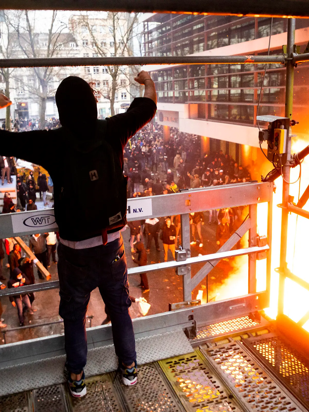 21.11.2021, Belgien, Brüssel: Ein Protestteilnehmer steht auf einem Baugerüst und gestikuliert in Richtung Demonstranten, die auf einer Straße vor einem Feuer stehen. Nach Polizeiangaben sind in Brüssel rund 35.000 Personen auf die Straße gegangen, um gegen die Coronamaßnahmen der Regierung zu demonstrieren. Bei Zusammenstößen musste die Polizei Wasserwerfer und Tränengas einsetzen. Foto: Hatim Kaghat/BELGA/dpa +++ dpa-Bildfunk +++