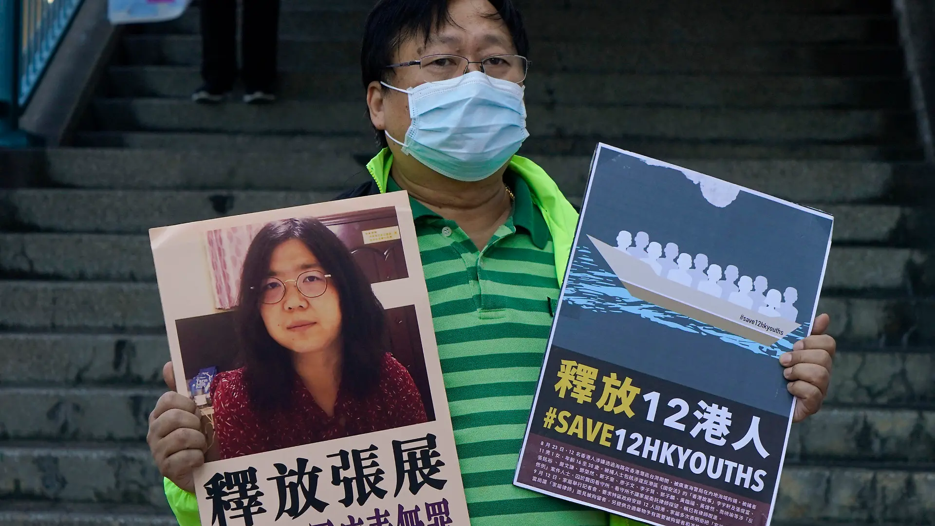 28.12.2020, China, Hongkong: Ein pro-demokratischer Aktivist zeigt zwei Plakate vor dem Verbindungsbüro der chinesischen Zentralregierung. Auf einem Plakat ist ein Foto der chinesischen Journalistin Zhang Zhan zu sehen. Zhang, eine ehemalige Anwältin und Journalistin aus Shanghai, wurde wegen ihrer Berichterstattung über den ersten Ausbruch des Coronavirus in Wuhan zu vier Jahren Gefängnis verurteilt. Die Aktivisten fordern die Freilassung von Zhang, sowie der 12 Hongkonger Aktivisten, die von den chinesischen Behörden auf See festgehalten werden. Foto: Kin Cheung/AP/dpa +++ dpa-Bildfunk +++