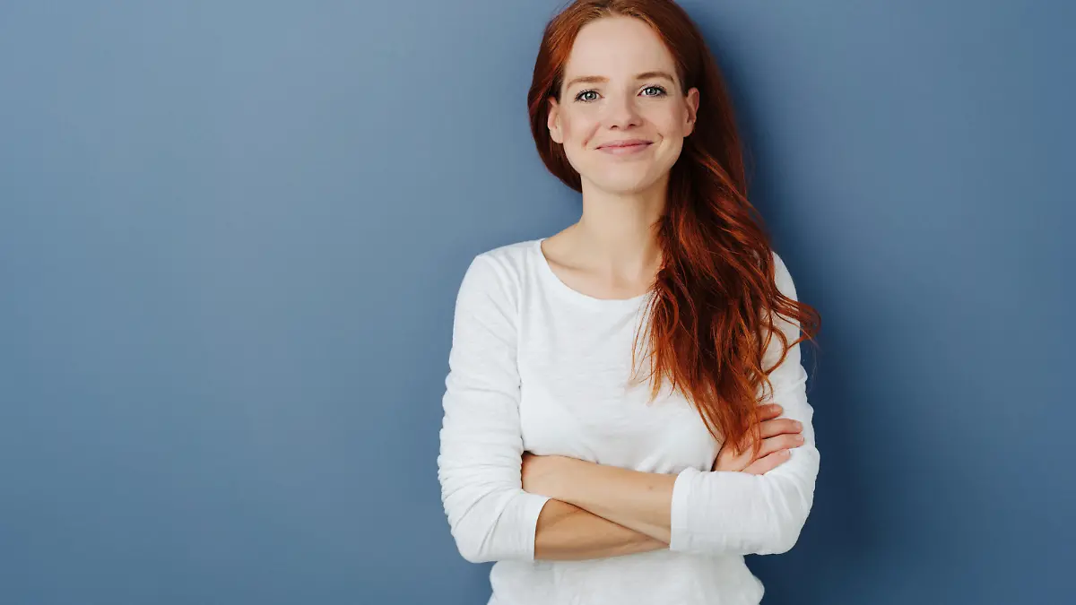Pleased confident young redhead woman with a beaming smile and folded arms posing on a blue studio background with copy space grinning at the camera