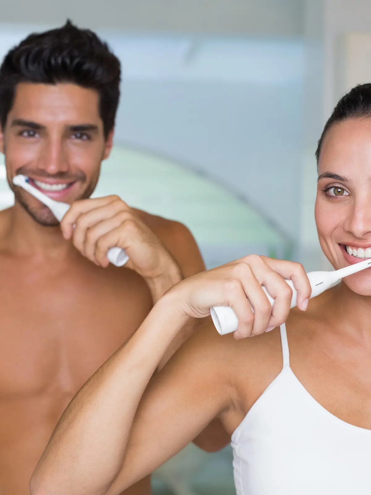 Attractive couple smile while using electric toothbrushes at home in bathroom