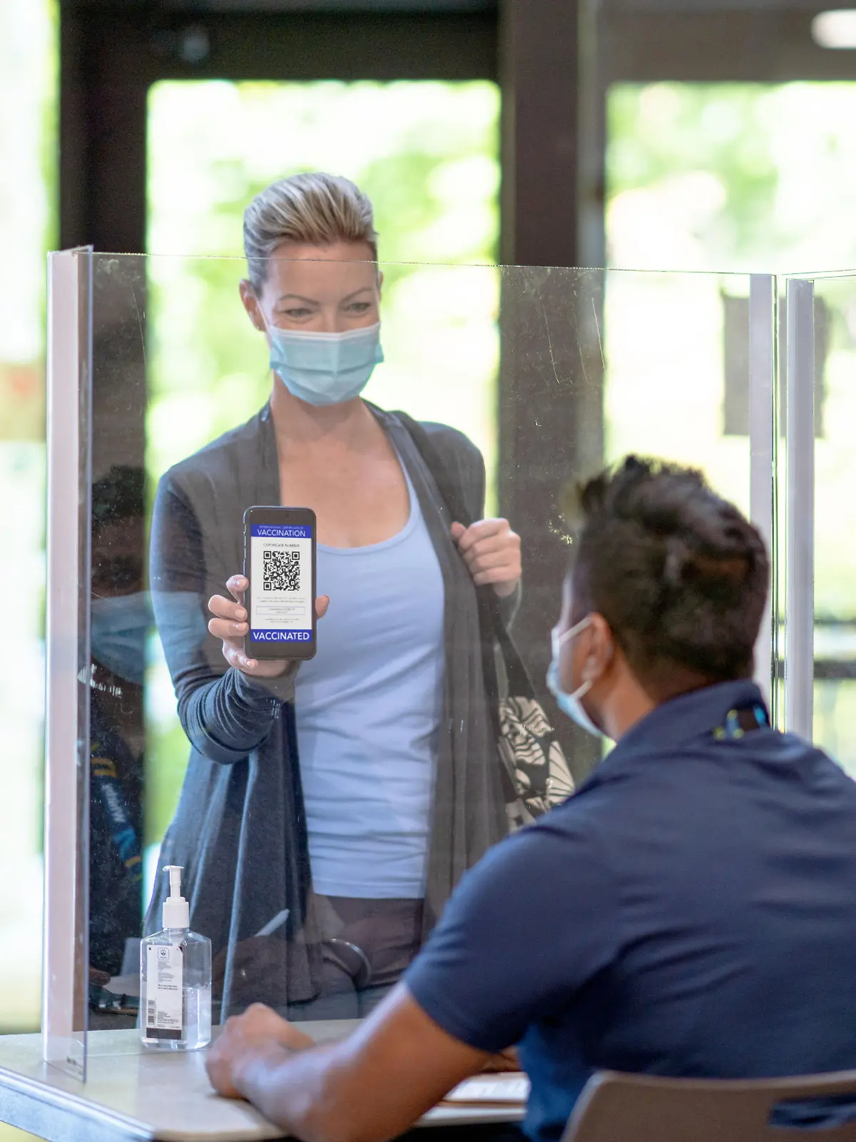 A middle aged woman enters a community centre and shows the attendant her vaccine passport on her cell phone.  She is dressed casually with her purse over her shoulder.  Both the woman and the attendant are wearing medical masks to protect them from COVID.