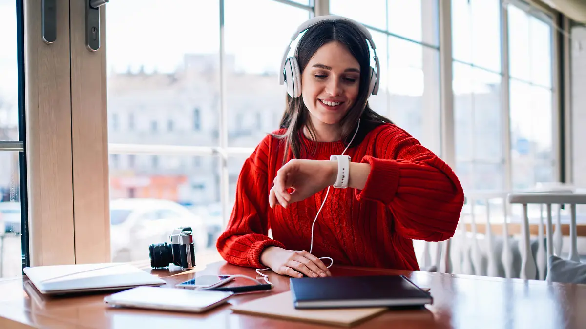Cheerful hipster girl listening new bestseller audiobook using application and earphones sitting at coffee shop, positive teenager enjoying funny playlist and checking time on wearable smartwatch