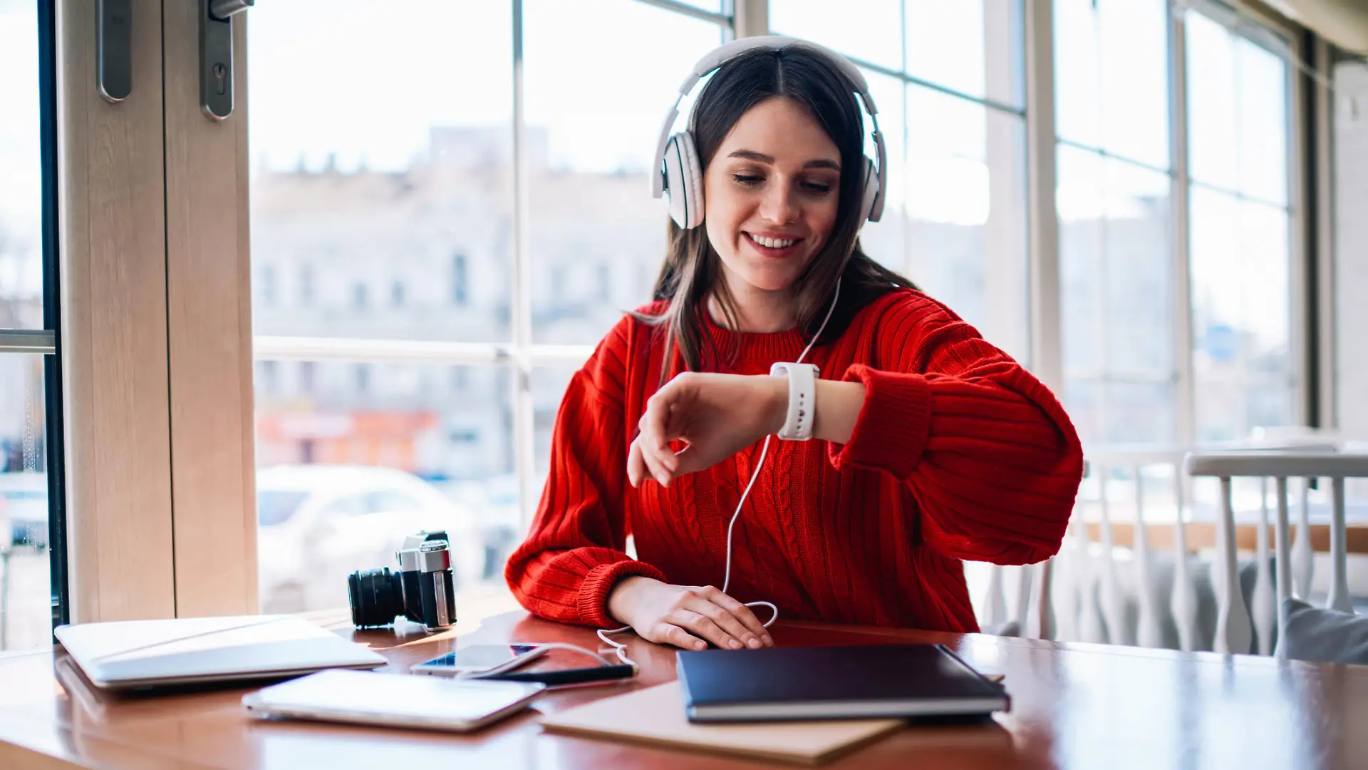 Cheerful hipster girl listening new bestseller audiobook using application and earphones sitting at coffee shop, positive teenager enjoying funny playlist and checking time on wearable smartwatch