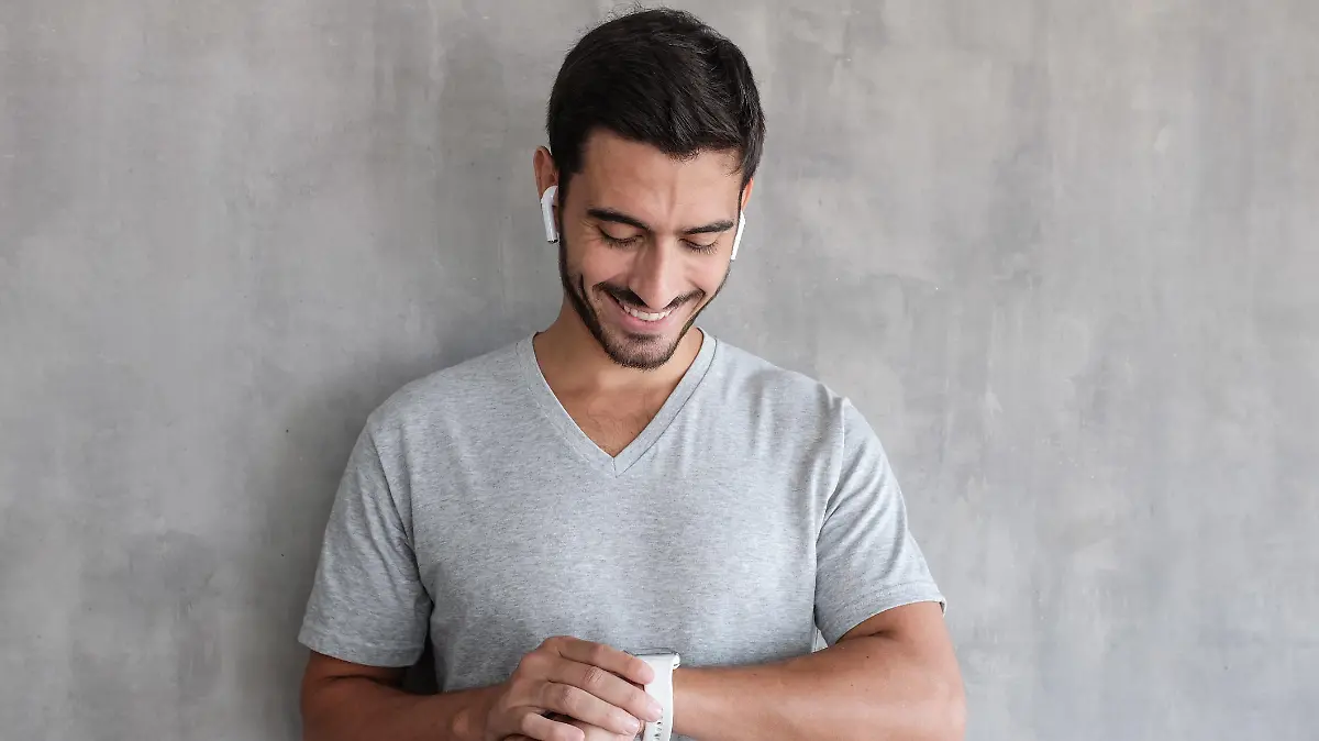 Young handsome man wearing wireless headphones and t shirt, checking smart watches with touch screen, standing against gray textured wall