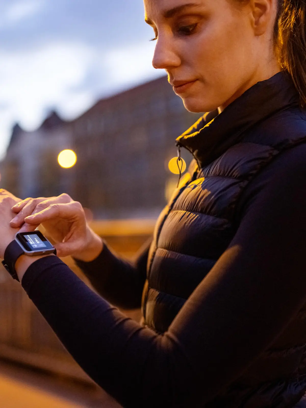 Young sporty woman checking smartwatch after running workout in the evening. Female in sportswear having a break standing outdoors after jogging in the city.