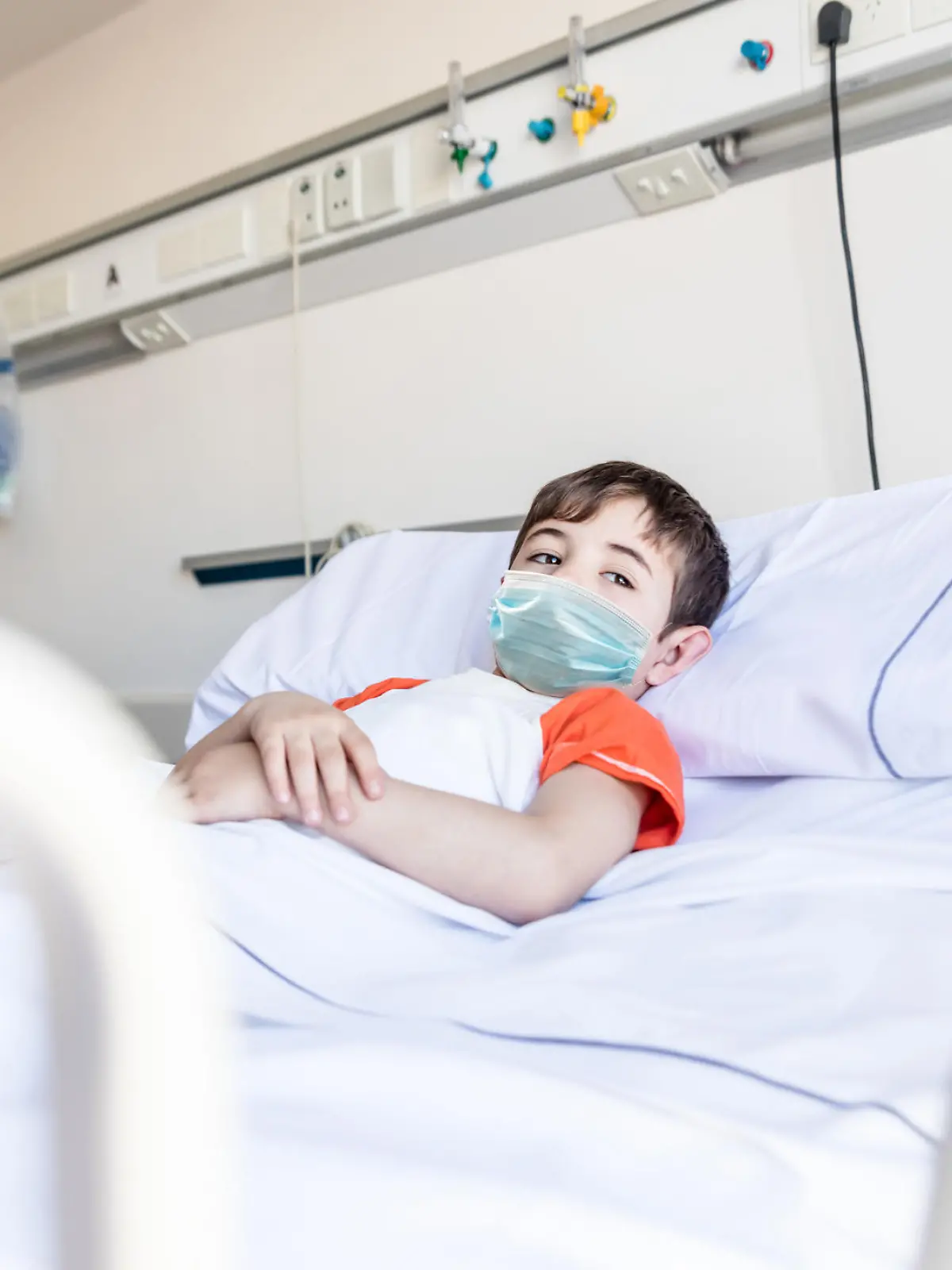 Pensive little boy lying down on hospital bed looking away with sadness wearing a protective facemask - Pandemic lifestyles