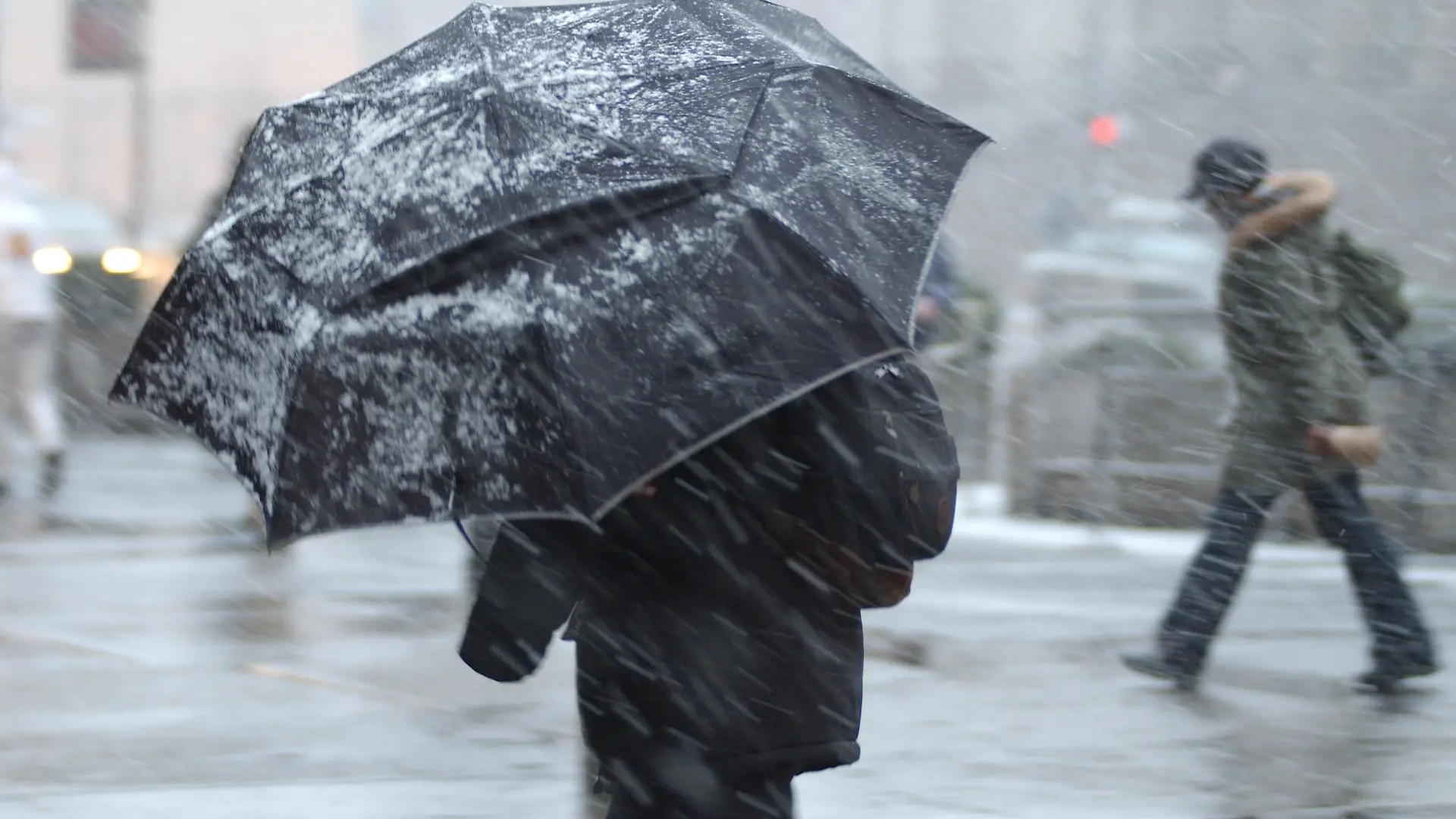 This is a photograph of a pedestrian in a snow storm with an aumbrella with a person in the background without an umbrella.