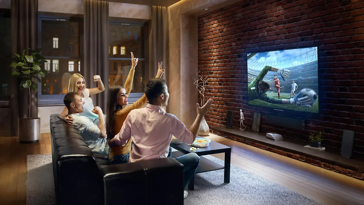 Two young couples cheering and watching Soccer game on TV. They are sitting on a sofa in the modern living room. The TV set is on the loft brick wall. It is evening outside the window.