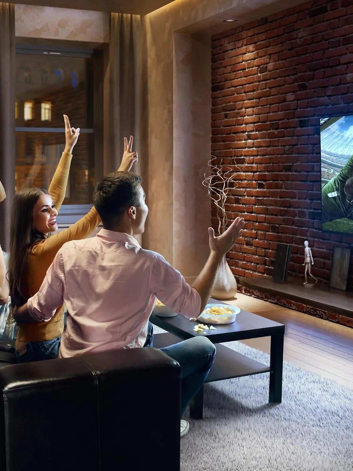 Two young couples cheering and watching Soccer game on TV. They are sitting on a sofa in the modern living room. The TV set is on the loft brick wall. It is evening outside the window.