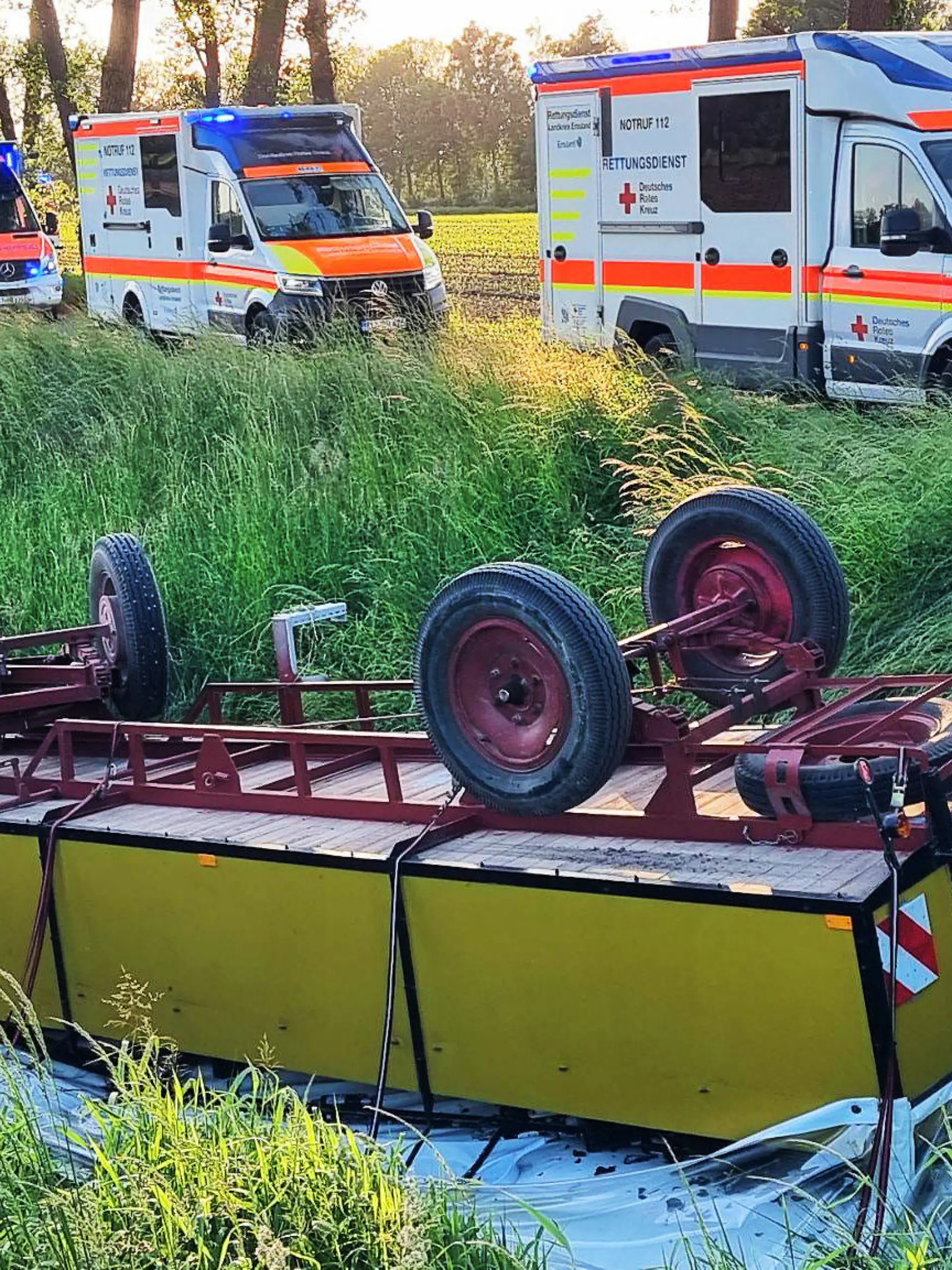 28.05.2023, Niedersachsen, Andervenne: Krankenwagen stehen an der Unfallstelle, an der im Emsland ein mit Ausflüglern besetzter Planwagen in einen Graben gestürzt war. Bei dem Unfall mit dem Planwagen sind zwei Frauen im Alter von 19 und 53 Jahren sowie ein 58-jähriger Mann bei Andervenne (Landkreis Emsland) schwer verletzt worden. Neun weitere Mitfahrende zwischen 18 und 82 Jahren kamen mit leichten Verletzungen davon, wie die Polizei mitteilte. Die Gruppe saß am Sonntagabend während eines Familienausflugs in dem Planwagen, der von einem Traktor gezogen wurde, als die Verbindung zwischen beiden Fahrzeugen riss. Der Planwagen rutschte vom Schotterweg in einen Graben und kam dort auf dem Dach zum Liegen. Grund für den Unfall war vermutlich eine gerissene Schweißnaht. Die Verletzten kamen in ein Krankenhaus. Foto: -/NWM-TV/dpa +++ dpa-Bildfunk +++