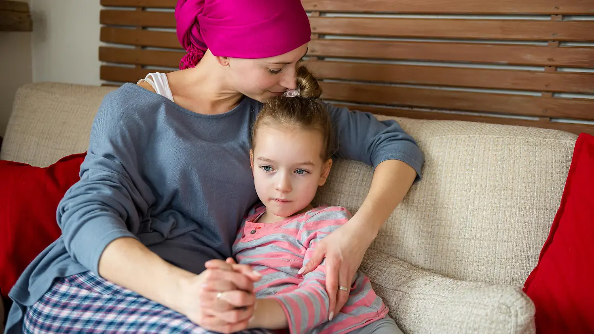 Young adult female cancer patient spending time with her daughter at home, relaxing on the couch. Cancer and family support concept.