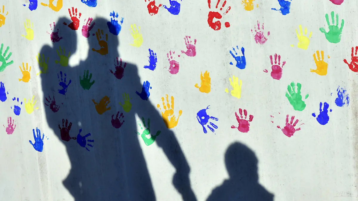 ARCHIV - 08.04.2015, Bayern, München: Ein Mann mit einem Kind auf dem Arm und einem an der Hand wirft einen Schatten auf eine mit bunten Handabdrücken bemalte Wand einer Kindertagesstätte. (zu dpa «Der Corona-Doppeljob trifft Kita-Eltern hart - und Mütter besonders?») Foto: Peter Kneffel/dpa +++ dpa-Bildfunk +++